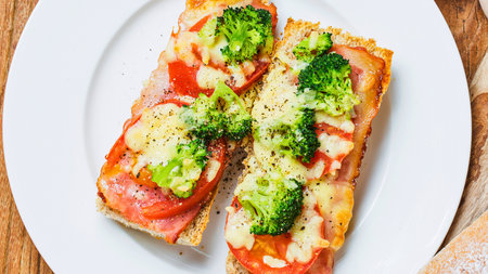 Two open faced sandwiches topped with melted cheese, tomato slices, and fresh broccoli florets, served on a white plate.の写真素材