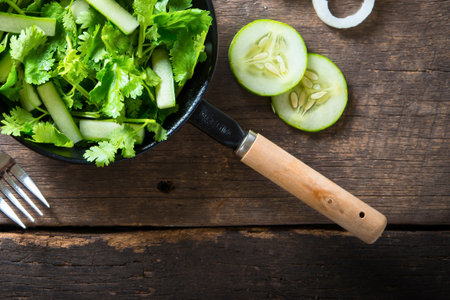 A vibrant green salad with cucumber slices and a fork on a textured wooden surface, ready to be served.の写真素材