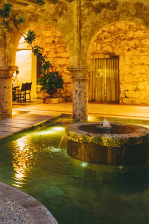 A tranquil courtyard at night, featuring a circular fountain with gentle water flow, surrounded by ancient stone architecture and warm ambient lighting.の写真素材