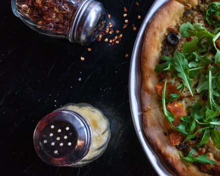 A close-up shot of a pizza with fresh green toppings and spices being sprinkled from shakers onto a dark surface.の写真素材