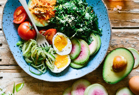 A colorful and nutritious breakfast bowl featuring avocado, soft-boiled eggs, greens, tomatoes, and zucchini noodles on a rustic wooden surface.の写真素材