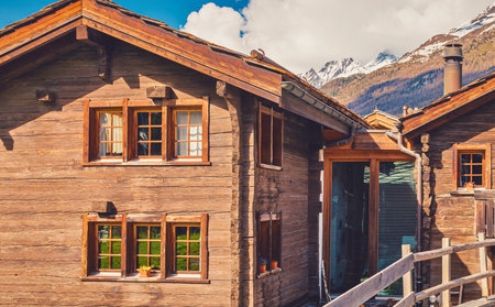 Traditional wooden chalets with rustic charm stand against a backdrop of majestic, snow-capped mountains under a bright blue sky.の写真素材
