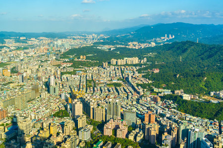 An aerial view captures a dense urban sprawl transitioning into verdant, tree-covered mountains under a bright, sunny sky.の写真素材