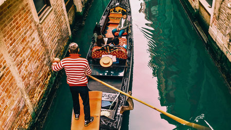 A gondolier in a striped shirt propels a gondola filled with passengers through a tight Venetian canal.の写真素材