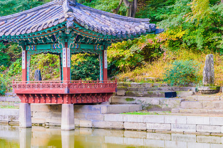A vibrant, multi-tiered Korean pavilion with red accents stands beside a calm body of water, surrounded by colorful autumn foliage.の写真素材