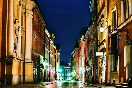 A narrow cobblestone street lined with historic buildings glows with warm lights under a dark night sky. The wet pavement reflects the vibrant illumination.の写真素材