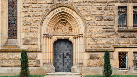 A grand stone building features a detailed Gothic archway above a dark metal door, flanked by two slender cypress trees.の写真素材