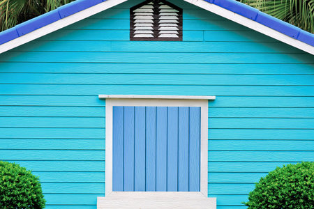 A vibrant blue wooden beach hut with a white framed door and window, topped by a blue roof and green foliage.の写真素材