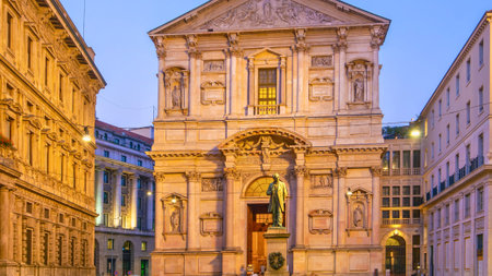 A grand, ornate Baroque church facade stands between tall buildings, illuminated by warm lights as dusk settles over the city.の写真素材