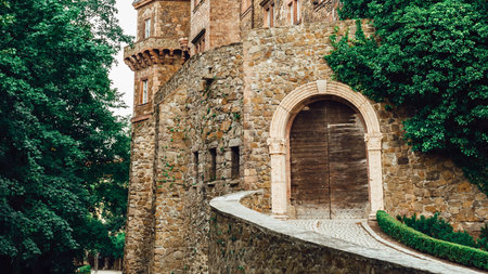 A historic stone castle features a grand arched doorway and a curved walkway lined with lush greenery.の写真素材