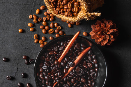 A dark, moody still life featuring roasted coffee beans spilling from a burlap sack, with cinnamon sticks and a pinecone on a textured surface.の写真素材