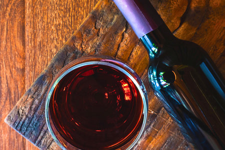 A close-up overhead view shows a full glass of deep red wine next to a dark wine bottle on a textured wooden table.の写真素材