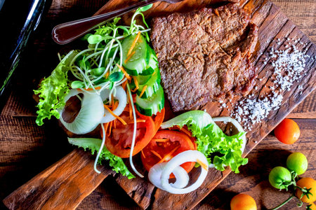 A rustic wooden board displays a thick slice of whole grain bread adorned with vibrant lettuce, tomato, onion, and sprouts. Small tomatoes are scattered nearby.の写真素材