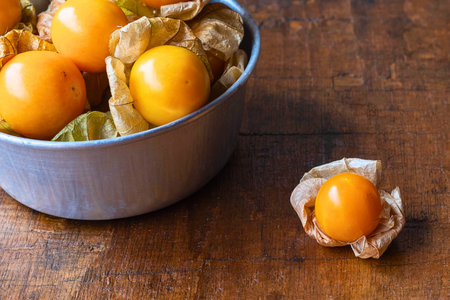 A close-up shot of bright orange gooseberries, some in a metal bowl, with one berry in its husk on a textured wooden table.の写真素材