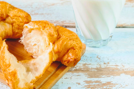 A close-up shot of a flaky, golden croissant torn open, next to a glass of white milk on a weathered wooden surface.の写真素材