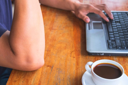 A person's arm and hand are visible as they type on a laptop. A cup of coffee sits nearby on a wooden surface.の写真素材