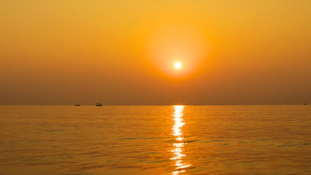 A brilliant orange sun sets over a tranquil sea, casting a shimmering golden path across the water. A small boat is visible on the horizon.の写真素材
