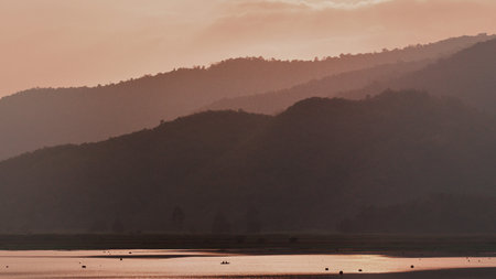 Layered mountain ranges fade into the distance under a soft, warm sunset sky, reflecting on a tranquil body of water.の写真素材