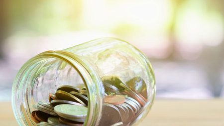 A close-up shot of a glass jar overflowing with coins, with sunlight glinting off the metal and the background softly blurred.の写真素材