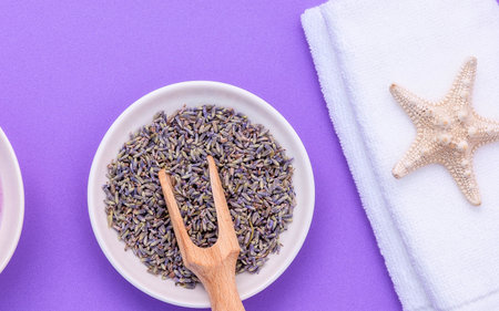 A top-down view of dried lavender flowers in a white oval bowl, a small wooden spoon, and a starfish on a purple background.の写真素材