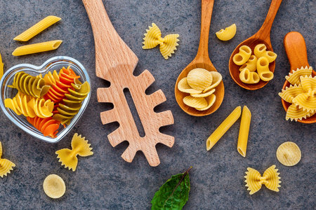A vibrant collection of various dried pasta shapes, including fusilli and farfalle, artfully arranged with wooden spoons and lemon slices on a textured dark background.の写真素材