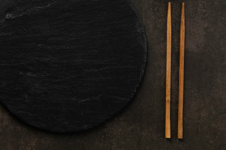 A dark, textured surface with a round black plate and two wooden chopsticks. Overhead shot with a minimalist aesthetic.の写真素材