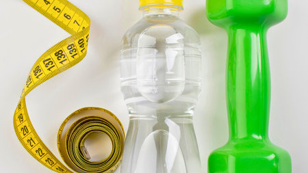 A yellow measuring tape, a bottle of water, and a green dumbbell are arranged on a white surface, symbolizing health and fitness.の写真素材