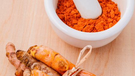 Fresh turmeric rhizomes and vibrant orange turmeric powder are displayed in a mortar on a wooden surface.の写真素材