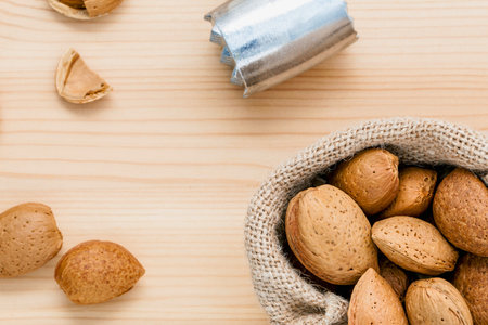 A rustic overhead shot shows a burlap sack overflowing with almonds, with a few scattered around on a light wooden background.の写真素材