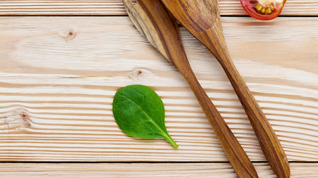 A close-up overhead view of two wooden spoons, a single spinach leaf, and a slice of tomato on a weathered wooden surface.の写真素材