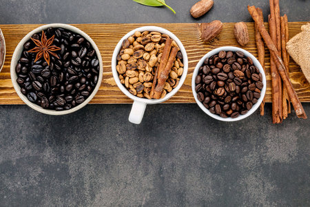 Overhead view of three white cups filled with different coffee beans alongside cinnamon sticks and star anise on a wooden board.の写真素材