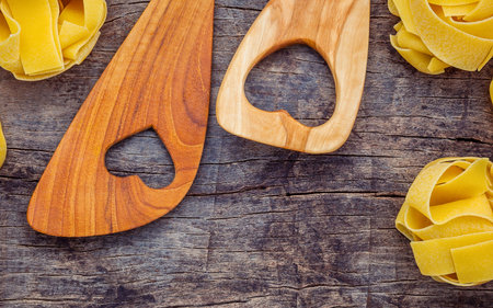 Two wooden spoons with heart cutouts rest beside nests of tagliatelle pasta on a textured, dark wooden surface.の写真素材