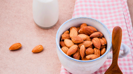 A white mug filled with almonds sits next to a bottle of milk and a wooden spoon on a pink checkered cloth.の写真素材