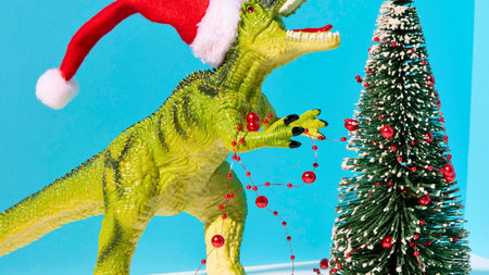 A green toy Tyrannosaurus Rex wearing a red Santa hat stands beside a small decorated Christmas tree, celebrating the holidays.の写真素材