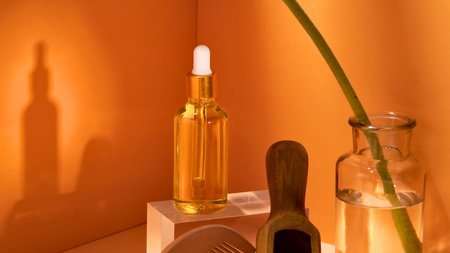 A glass dropper bottle filled with golden liquid stands on a pedestal, casting a long shadow against an orange wall. A plant stem is visible.の写真素材