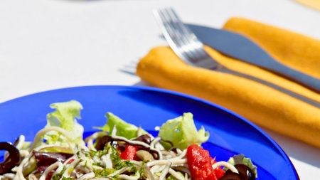 A close-up of a colorful salad featuring lettuce, olives, and other vegetables served on a bright blue plate with cutlery in the background.の写真素材