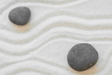 Two dark, smooth stones rest on a bed of white sand meticulously raked into gentle waves, creating a tranquil and minimalist scene.の写真素材