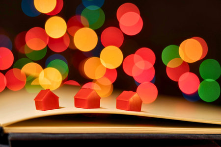Small red house miniatures are placed on an open book, with blurred colorful lights creating a magical bokeh effect in the background.の写真素材