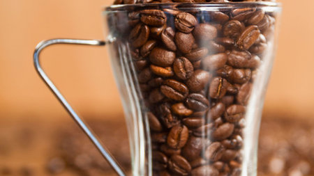 A close-up shot of a clear glass mug overflowing with dark roasted coffee beans, set against a blurred background of more beans.の写真素材