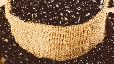 A textured burlap sack is filled to the brim with dark, glossy black beans, creating an appealing food background.の写真素材