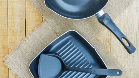 A top-down view of a frying pan, grill pan, and spatula arranged on a rustic wooden surface with...
