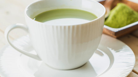 A close-up of a frothy matcha latte in a ribbed white mug, with a small dish of matcha powder in the background.の写真素材