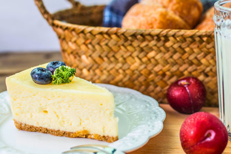 A creamy slice of cheesecake topped with blueberries sits on a plate, with plums and a basket in the...