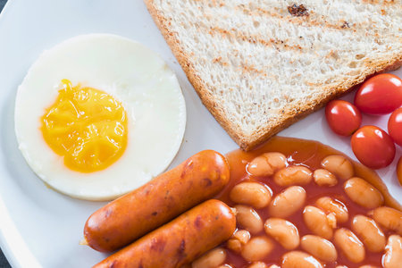 A close-up overhead view of a delicious breakfast featuring sausages, baked beans, toast, cherry...