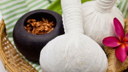 A close-up of herbal compress balls and a dark bowl filled with dried herbs, alongside a vibrant pink flower, suggesting a spa or massage setting.の写真素材