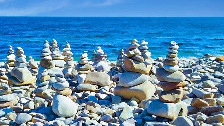 Numerous carefully balanced rock stacks stand on a pebble beach, with the blue ocean and sky in the...
