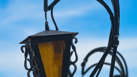 A dark, wrought-iron lantern with a yellow glass pane hangs from a decorative metal pole against a backdrop of a pale blue sky with wispy clouds.の写真素材