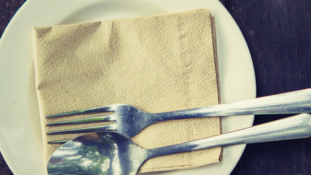 A clean white plate holds a folded brown napkin with a fork and spoon resting on top, ready for a meal.の写真素材