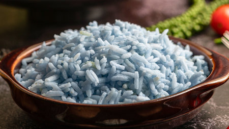 A close-up of fluffy, light blue rice piled high in a brown ceramic bowl, with a hint of greenery and a red tomato in the background.の写真素材