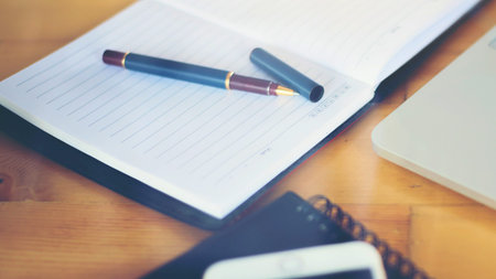 A close-up shot of an open notebook with a sophisticated pen, a smartphone, and a laptop on a wooden...